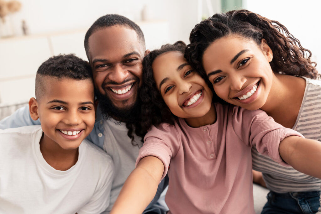 Capturing Moments. Portrait of happy loving black family of four people taking selfie together, closeup. Positive parents posing with their kids and smiling, girl holding camera, pov
